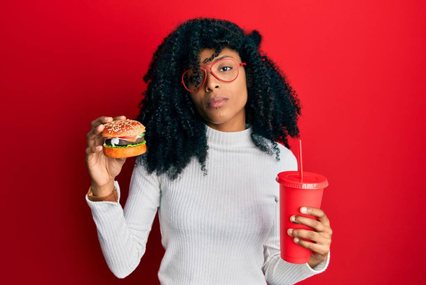 Afro-Amerikaanse vrouw met afrohaar die een lekkere klassieke hamburger en frisdrank eet ontspannen met serieuze uitdrukking op haar gezicht. eenvoudig en natuurlijk kijkend naar de camera.  - Foto, afbeelding