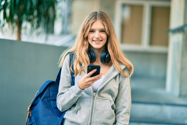 piękny biały student nastolatek uśmiecha się szczęśliwy za pomocą smartfona na uniwersytecie. - Zdjęcie, obraz