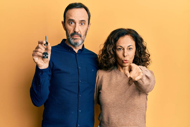 Hispánský pár středního věku drží klíče nového domova ukazující prstem na fotoaparát a na vás, sebevědomé gesto vypadá vážně  - Fotografie, Obrázek