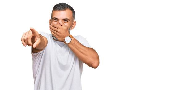 Middelbare leeftijd knappe man draagt casual witte tshirt lachen naar je, wijzende vinger naar de camera met hand over mond, schaamte uitdrukking  - Foto, afbeelding