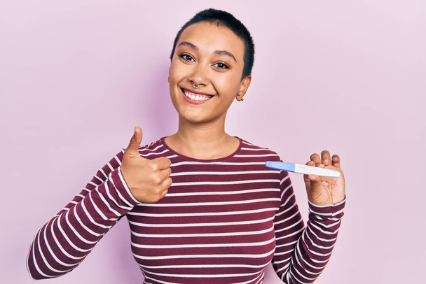 Beautiful hispanic woman with short hair holding pregnancy test result smiling happy and positive, thumb up doing excellent and approval sign  - Photo, Image