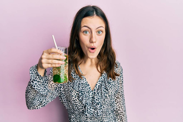 Young brunette woman drinking mojito glass scared and amazed with open mouth for surprise, disbelief face  - Фото, изображение