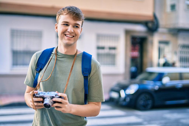 Jeune homme touristique caucasien souriant heureux en utilisant une caméra vintage à la ville. - Photo, image