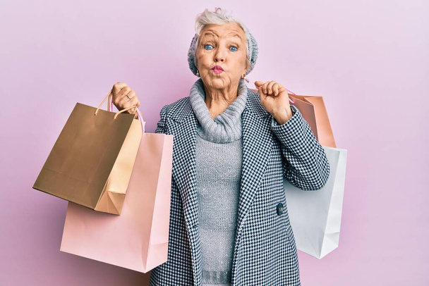 Senior grey-haired woman holding shopping bags puffing cheeks with funny face. mouth inflated with air, catching air.  - Foto, imagen