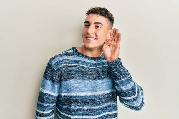 Ισπανόφωνος νεαρός άνδρας που φοράει ένα απλό χειμωνιάτικο πουλόβερ χαμογελώντας με το χέρι πάνω από το αυτί ακούει και ακούει φήμες ή κουτσομπολιά. έννοια της κώφωσης.  - Φωτογραφία, εικόνα