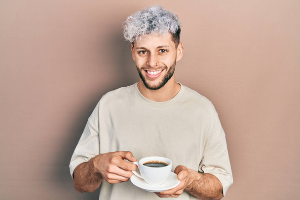 Młody Latynos z nowoczesnymi farbowanymi włosami pijący filiżankę kawy uśmiechnięty ze szczęśliwym i chłodnym uśmiechem na twarzy. wykazujące zęby.  - Zdjęcie, obraz