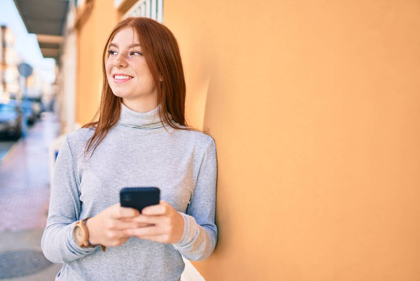 Jovem irlandês adolescente sorrindo feliz usando smartphone na cidade. - Foto, Imagem