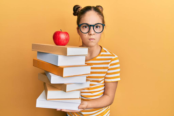 Gyönyörű barna kislány kezében egy halom könyvet egy almával a tetején szkeptikus és ideges, homlokráncolás ideges, mert a probléma. negatív személy.  - Fotó, kép