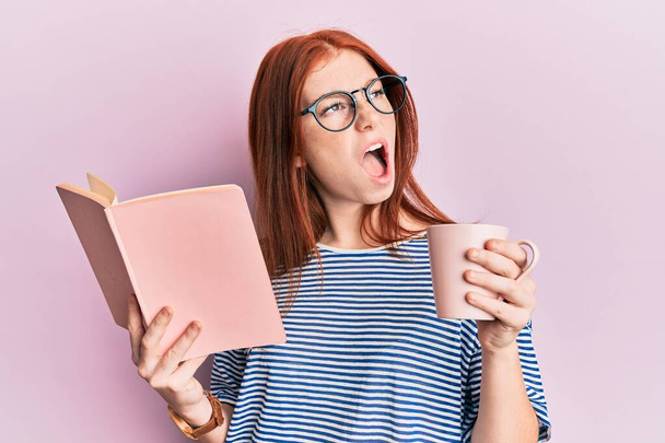 Young red head girl reading a book and drinking a cup of coffee angry and mad screaming frustrated and furious, shouting with anger looking up.  - Photo, Image