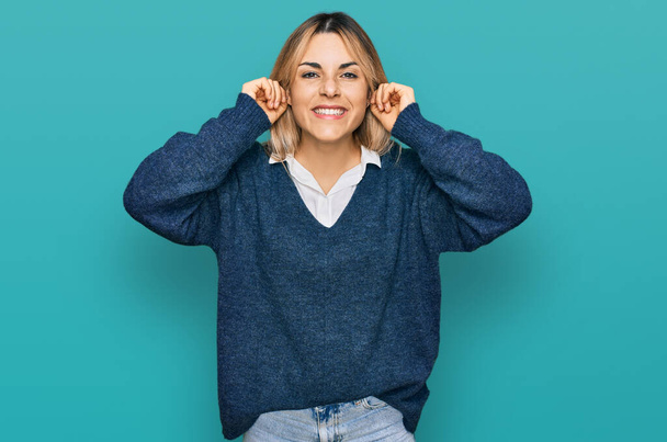 Mujer caucásica joven con ropa casual sonriendo tirando de las orejas con los dedos, gesto divertido. problema de audición  - Foto, Imagen