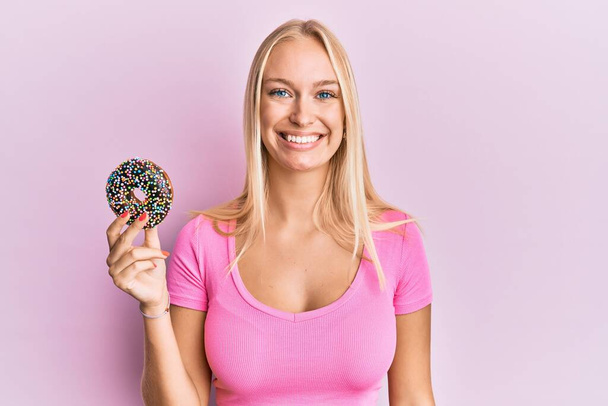 Young blonde girl holding donut looking positive and happy standing and smiling with a confident smile showing teeth  - Photo, Image