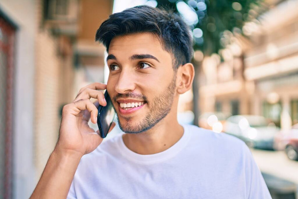 Genç Latin adam gülümsüyor ve şehirde akıllı telefondan konuşuyor.. - Fotoğraf, Görsel