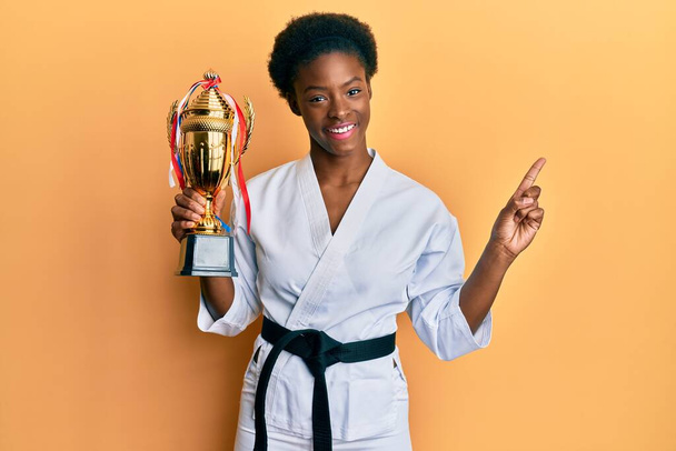 Giovane ragazza afroamericana con indosso karate kimono tenendo trofeo sorridente felice indicando con mano e dito di lato  - Foto, immagini