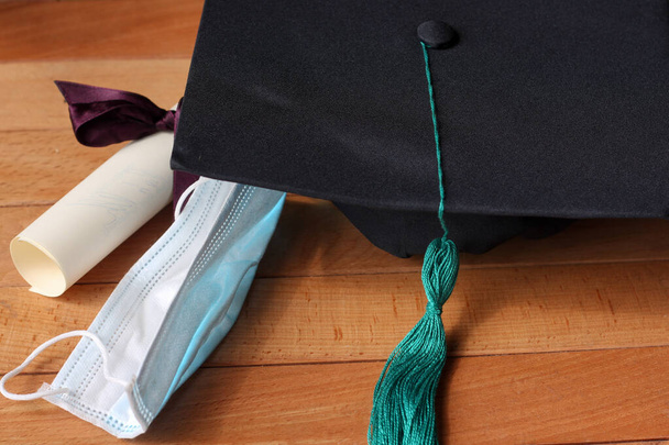 Absolventenhut und Diplom auf einem hölzernen Hintergrund zusammen mit einer medizinischen Schutzmaske - Foto, Bild