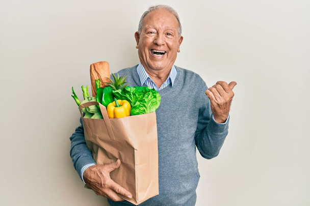 Senior caucasian man holding paper bag with bread and groceries pointing thumb up to the side smiling happy with open mouth  - Photo, Image