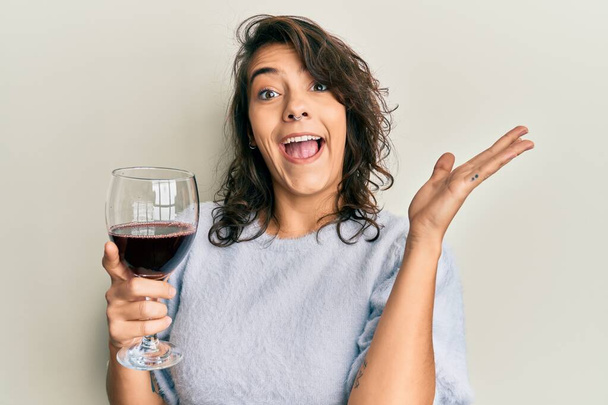 Jovem hispânica bebendo um copo de vinho tinto celebrando a vitória com sorriso feliz e expressão vencedora com as mãos levantadas  - Foto, Imagem