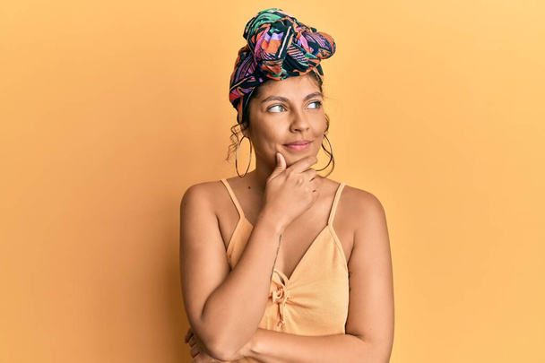 Young hispanic girl wearing hair turban over yellow background with hand on chin thinking about question, pensive expression. smiling with thoughtful face. doubt concept.  - Фото, зображення