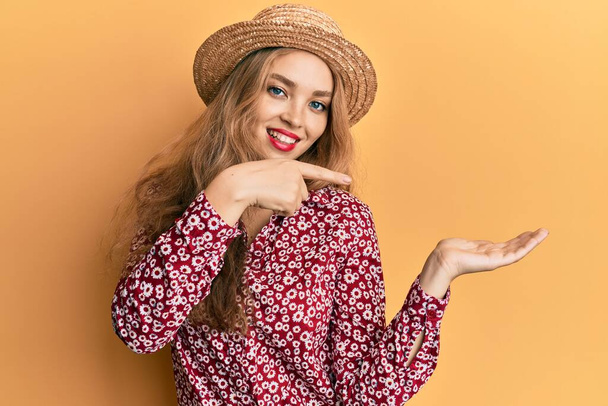 Piękna blondynka kaukaska kobieta w letniej czapce zdumiony i uśmiechnięty do aparatu podczas prezentacji z ręki i wskazując palcem.  - Zdjęcie, obraz