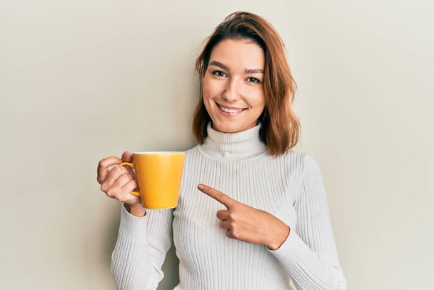 Młoda kaukaska kobieta trzyma kawę uśmiechając się szczęśliwy wskazując ręką i palcem  - Zdjęcie, obraz