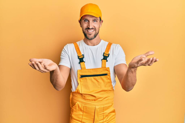 Young handsome man wearing handyman uniform over yellow background smiling cheerful offering hands giving assistance and acceptance.  - Foto, imagen
