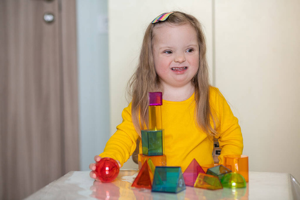 Piękna dziewczyna z zespołem Downa bawi się z konstruktorem, jasnymi, przezroczystymi postaciami, wczesnym rozwojem niepełnosprawnego dziecka. - Zdjęcie, obraz