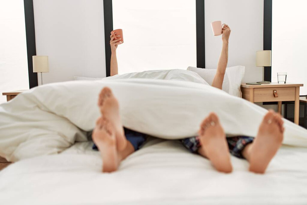 Un paio svegli sotto coperta. Facendo colazione tenendo una tazza di caffè sollevata sul letto. - Foto, immagini