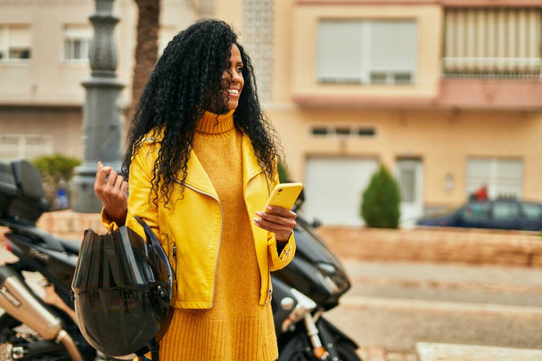 Μέση ηλικία Αφροαμερικανή γυναίκα κρατώντας κράνος μοτοσικλέτας και χρησιμοποιώντας smartphone στην πόλη. - Φωτογραφία, εικόνα