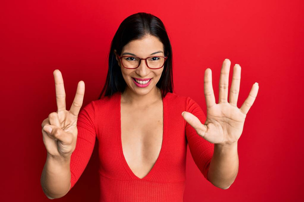 Mujer latina joven usando ropa casual y gafas que muestran y señalan con los dedos número siete mientras sonríe confiada y feliz.  - Foto, imagen