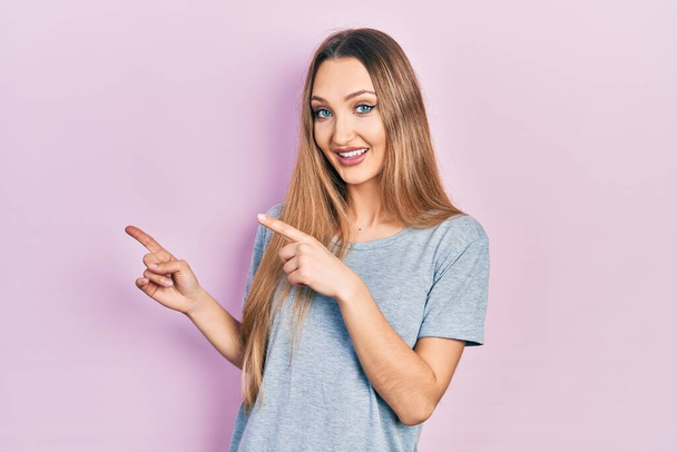 Mladá blondýna dívka na sobě ležérní tričko s úsměvem a při pohledu na kameru ukazuje dvěma rukama a prsty na stranu.  - Fotografie, Obrázek