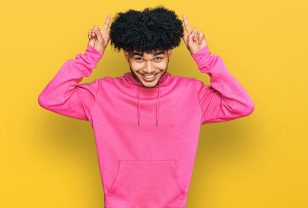 Junger afrikanisch-amerikanischer Mann mit Afrohaaren trägt lässiges rosafarbenes Sweatshirt und posiert lustig und verrückt mit den Fingern auf dem Kopf als Hasenohren, lächelnd fröhlich  - Foto, Bild