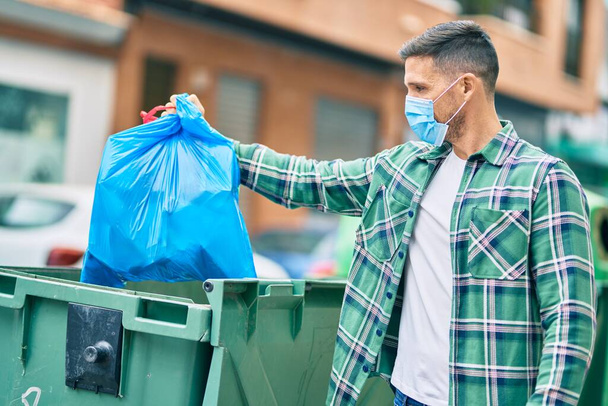 Joven hombre caucásico con máscara médica arrojando la bolsa de residuos al contenedor en la ciudad. - Foto, imagen