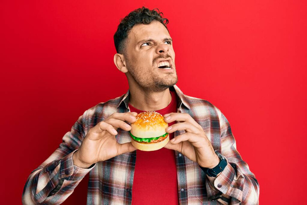 Nuori latinomies syö maukasta klassista hampurilaista vihaisena ja hulluna huutaen turhautuneena ja raivoissaan, huutaen vihasta. raivo ja aggressiivinen käsite.  - Valokuva, kuva