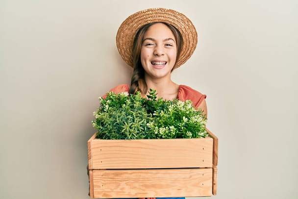 Beautiful brunette little girl wearing gardener hat holding wooden plant pot smiling and laughing hard out loud because funny crazy joke.  - Photo, Image