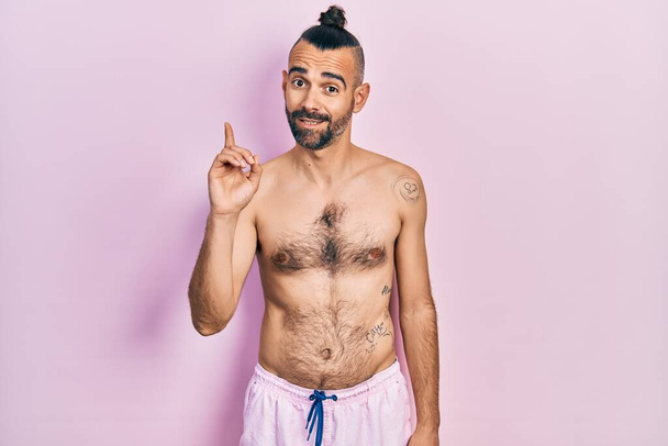 Fiatal spanyol férfi félmeztelen fürdőruhát visel, első számú ujjal mutogat, miközben magabiztosan és boldogan mosolyog..  - Fotó, kép