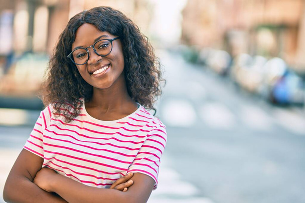 Joven afroamericana americana con los brazos cruzados sonriendo feliz de pie en la ciudad. - Foto, Imagen