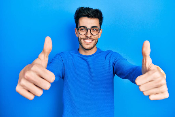 Молодой латиноамериканец в повседневной одежде и очках, одобряющий позитивный жест рукой, улыбается и радуется успеху. жест победителя.  - Фото, изображение