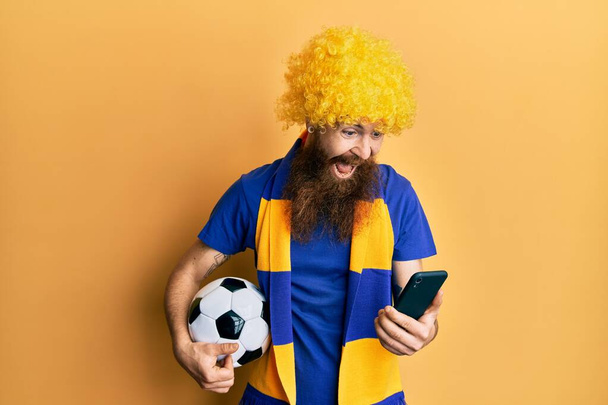 Homme rousse avec longue barbe football hooligan acclamations jeu tenant smartphone célébrant fou et étonné pour le succès avec les yeux ouverts hurlant excité.  - Photo, image