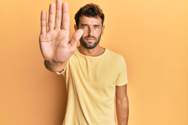 Pohledný muž s plnovousem na sobě nenucené žluté tričko přes žluté pozadí přestat zpívat s dlaní ruky. varovný výraz s negativním a vážným gestem na tváři.  - Fotografie, Obrázek