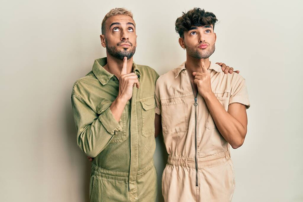 Homosexual pareja gay de pie juntos usando casual jumpsuit pensamiento concentrado acerca de la duda con el dedo en la barbilla y mirando hacia arriba preguntando  - Foto, Imagen