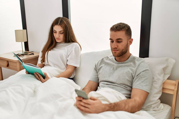 Junges kaukasisches Paar liest Buch und nutzt Smartphone im Bett. - Foto, Bild