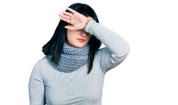 Młoda Latynoska plus kobieta w zimowym szaliku zakrywająca oczy ramieniem, wyglądająca poważnie i smutno. Koncepcja niewidoczności, ukrywania i odrzucania  - Zdjęcie, obraz