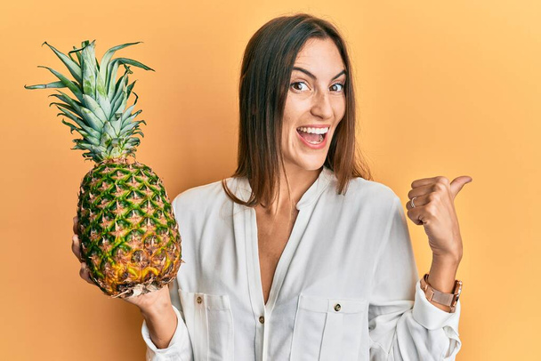 Młoda piękna kobieta trzyma ananasa wskazując kciuk w górę do boku uśmiechając się szczęśliwy z otwartymi ustami  - Zdjęcie, obraz