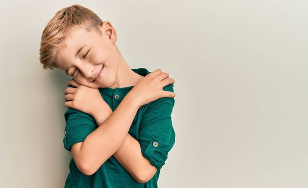 Kleiner kaukasischer Junge in lässiger Kleidung, der sich glücklich und positiv umarmt und selbstbewusst lächelt. Selbstliebe und Selbstfürsorge  - Foto, Bild