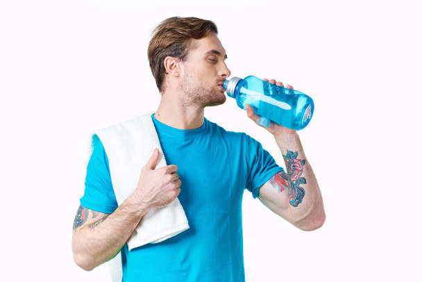 atleta hombre bebe agua de una botella sobre un fondo blanco y una toalla en su hombro - Foto, imagen
