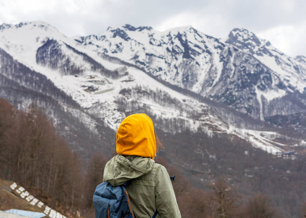 Mujer joven en capucha amarilla con una mochila por detrás disfrutando de la vista de los picos de montaña caucásicos cubiertos de nieve, senderismo, estilo de vida activo, paisaje - Foto, imagen