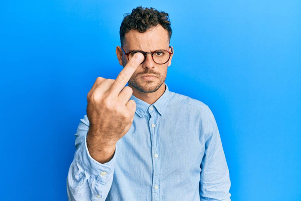 junger hispanischer Mann in lässiger Kleidung und Brille mit Mittelfinger, unhöflichem und unhöflichem Gesichtsausdruck  - Foto, Bild