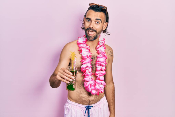 Fiatal spanyol férfi fürdőruhában és hawaii lejben, trópusi koktélt iszik, és vidáman kinyújtja a nyelvét. érzelem koncepció.  - Fotó, kép