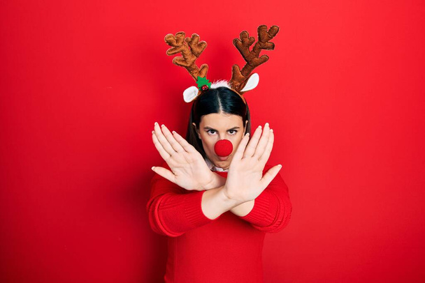 Young hispanic woman wearing deer christmas hat and red nose rejection expression crossing arms and palms doing negative sign, angry face  - Photo, Image
