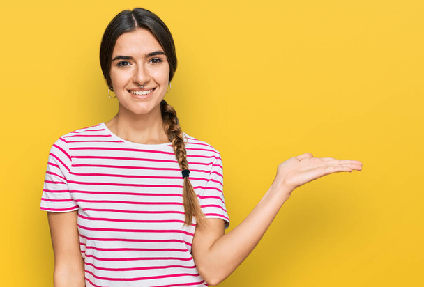 Mujer hispana joven vistiendo ropa casual sonriendo alegre presentando y señalando con la palma de la mano mirando a la cámara.  - Foto, Imagen