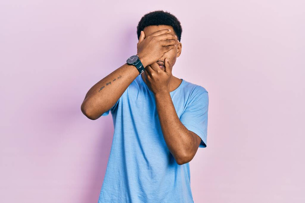 Genç Afro-Amerikalı bir adam günlük mavi tişört giyip gözlerini ve ağzını elleriyle kapatmış, şaşırmış ve şok olmuş. duygu gizleme  - Fotoğraf, Görsel